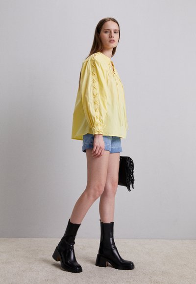Femme debout portant un haut à manches longues jaune avec des manches en broderie, un short en denim, des bottes à talons noires et tenant un sac à franges noir.