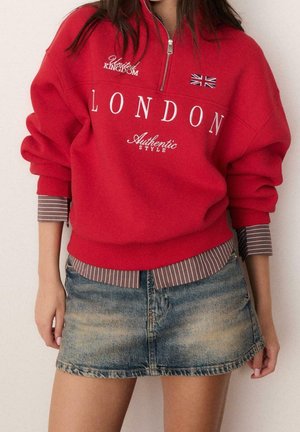 Osoba nosząca czerwoną bluzę z napisem "London" i flagą Wielkiej Brytanii, nałożoną na pasiastą koszulę, w połączeniu z wyblakłą jeansową mini spódniczką.