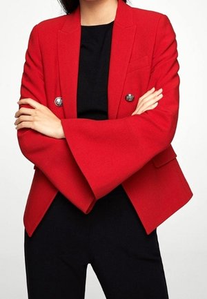 Kvinde iført en rød blazer med udvidede ærmer og sølvknapper, armene krydset, over en sort top og sorte bukser.