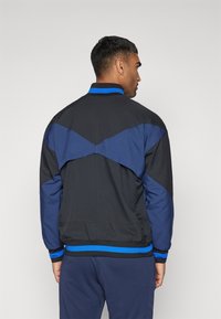 Giacca nera e blu con un alto colletto, strisce blu a contrasto e un dettaglio unico a yoke angolato sul retro. Tessuto di trama liscia.