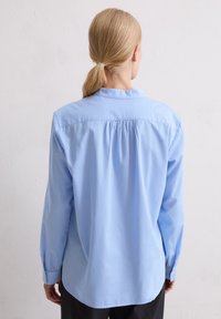 Osoba o blond włosach związanych w niski kucyk, ubrana w jasnoniebieską koszulę z długim rękawem, widziana od tyłu na tle jednolitej białej ściany.
