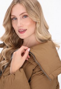 Jachetă pufos maro cu guler înalt, textură matlasată și detalii cu fermoar. Modelul atinge gulerul cu un fundal natural delicat.