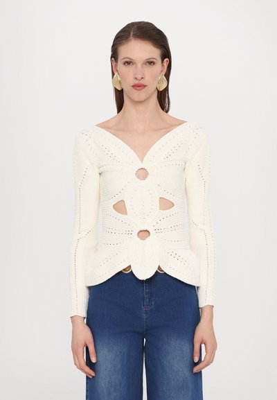 Haut en maille blanc à manches longues et col en V profond. Présente des découpes et un motif floral avec des sections ouvertes à la taille.