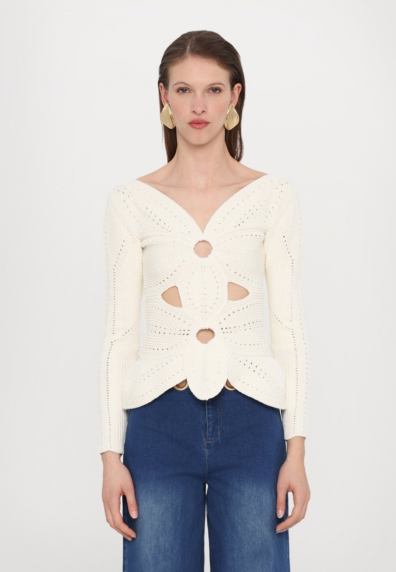 White knitted sweater with a deep V-neck, cut-out patterns, and textured detailing. Paired with blue wide-leg jeans.