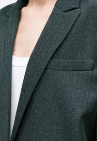 Personne portant un blazer en chevron gris foncé sur un haut blanc côtelé, montrant la clavicule et la partie supérieure de la poitrine.