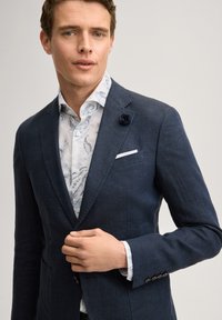 Homme portant un blazer bleu foncé avec une épingle florale à la boutonnière et une chemise blanche à motifs, ajustant le devant du blazer.