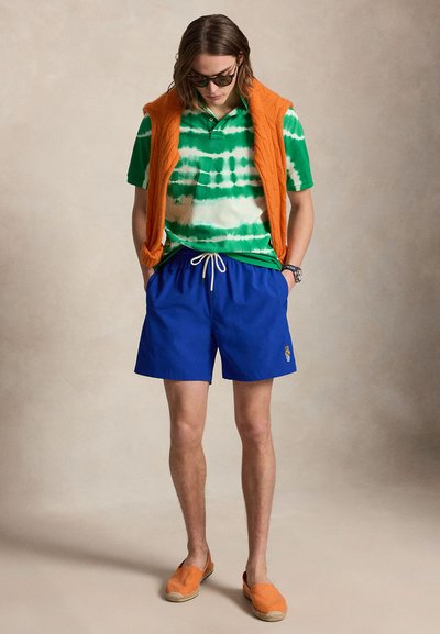 Νέος άνδρας που φοράει γυαλιά ηλίου, πορτοκαλί αμάνικο πουλόβερ πάνω από πράσινο μπλουζάκι με tie-dye, μπλε σορτς με κορδόνι και πορτοκαλί εσπαντρίγιες, στέκεται με τα χέρια στις τσέπες.