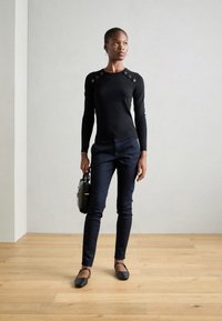 Černý svetr s dlouhými rukávy a dekorativními knoflíky na ramenech, spárovaný s tmavomodrými úzkými kalhotami a černými plochými botami, držící malou kabelku.