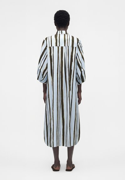 Άτομο που στέκεται με την πλάτη στον θεατή, φορώντας ένα μακρύ, φαρδύ φόρεμα με κάθετες καφέ και λευκές ρίγες και μαύρα σανδάλια.