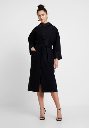 Zwarte wollen trenchcoat met een ceintuur in de taille, grote opgestikte zakken en brede manchetten. Heeft een gelapte kraag en een knielange ontwerp.