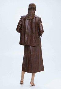 Ruskea nahkatakki, jossa on räätälöity malli ja pitkät hihat, yhdistettynä samanlaiseen pitkään hameeseen; molemmissa on sileä pinta ja siistit linjat.