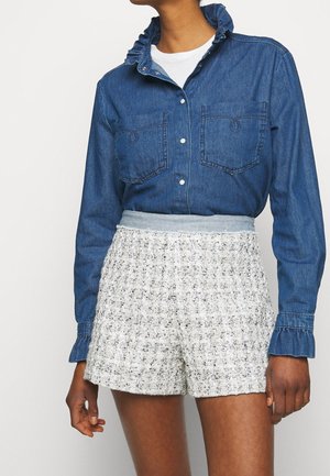 Personne portant une chemise en jean bleue boutonnée avec des manches et un col froncés, rentrée dans un short blanc texturé taille haute.