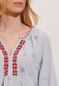 Jasnoniebieska bluzka z czerwonym i białym haftem kwiatowym wzdłuż dekoltu i klatki piersiowej. Miękka, teksturowana tkanina o luźnym kroju.