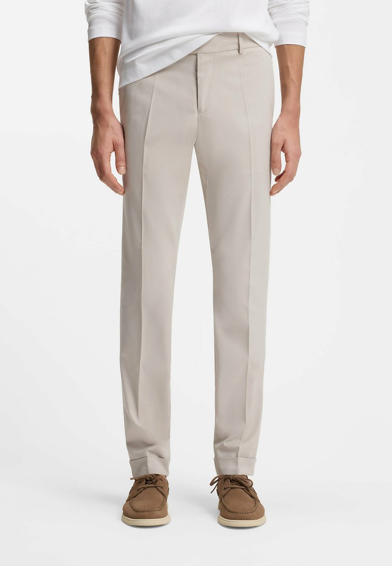 Homme portant un pantalon beige sur mesure, des chaussures en daim marron clair à lacets et une chemise blanche à manches longues, sur un fond blanc.