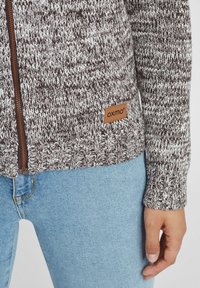 Brun og hvit strikket zip-genser med ribbestrikket nederkant og mansjetter, med et lærlogo-patch. Kombinert med lyseblå jeans.