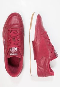 Červené kožené tenisky s texturovaným povrchem, rovnými tkaničkami a bílou gumovou podrážkou. Obsahují značku Reebok na jazyku a boku.