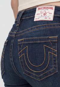 Close-up of dark blue True Religion jeans back pocket with yellow stitching and white brand label on waistband.