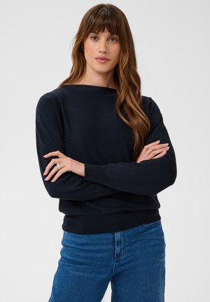 Femme aux longs cheveux bruns portant un pull noir et un jean bleu, debout les bras croisés devant un fond clair uni.