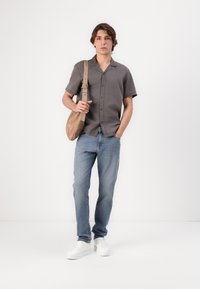Chemise gris à manches courtes avec des boutons, jean bleu clair, baskets blanches et un sac à épaule beige. Tissu uni avec une coupe décontractée.