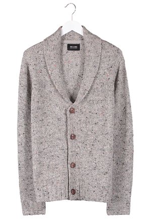 Mand iført grå knappet cardigan, olivengrøn skjorte, sorte bukser, mørkerøde sneakers og brune skulderremme, stående med hænderne i lommerne.