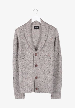 Uomo che indossa un cardigan grigio abbottonato, camicia oliva, pantaloni neri, scarpe da ginnastica marroni e bretelle marroni, in piedi con le mani in tasca.