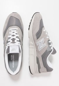 Szare zamszowe i siateczkowe sneakersy z białymi sznurówkami i akcentami. Oferują wyściełany kołnierz, gumową podeszwę oraz odblaskowe logo "N" po bokach.