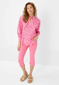 Blusa rosa con design a occhielli e dettaglio a fiocco, abbinata a pantaloni cropped coordinati e sandali multicolor con fibbie decorative.