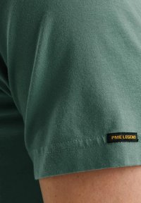 T-shirt in cotone verde con maniche corte, caratterizzata da un'etichetta nera con testo giallo sull'orlo. Texture liscia con un design semplice.