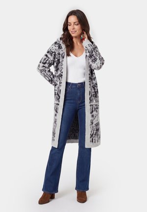 Kvinde iført en lang grå og sort mønstret cardigan, hvid top, blå jeans, brune støvler og farverige øreringe, der står med den ene hånd på nakken.