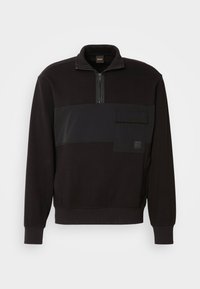 Felpa nera con zip a metà colletto, un ampio pannello orizzontale sulla parte anteriore e una tasca frontale con una patch del logo.