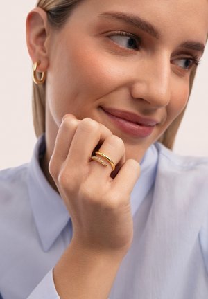 Anillos de oro en una mano; uno es una banda lisa y el otro presenta piedras preciosas de colores incrustadas en un diseño delgado. Camisa azul claro con cuello.