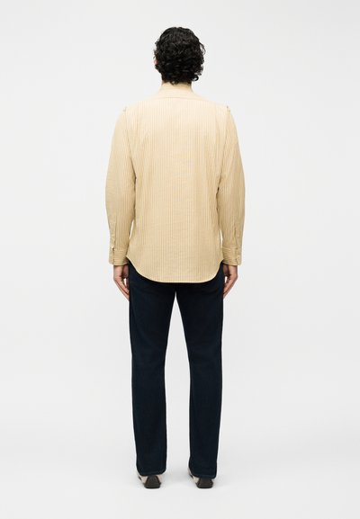 Άνδρας με σγουρά μαλλιά που φοράει μπεζ ριγέ πουκάμισο με μακριά μανίκια και σκούρο παντελόνι, στέκεται γυρισμένος προς τα πίσω μπροστά σε ένα απλό φόντο.