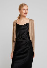 Käkiline kuldne lühike kardigan pikkade varrukatega, mille all on must satin kleit, millel on südamekujuline dekoltee ja must vöö.