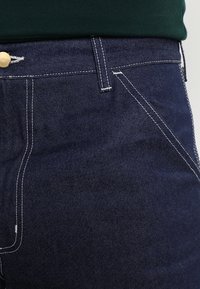 Mörkblå denimjeans har en klassisk femfickedesign med vita sömmar och en guldknaff vid midjan. Slät struktur.
