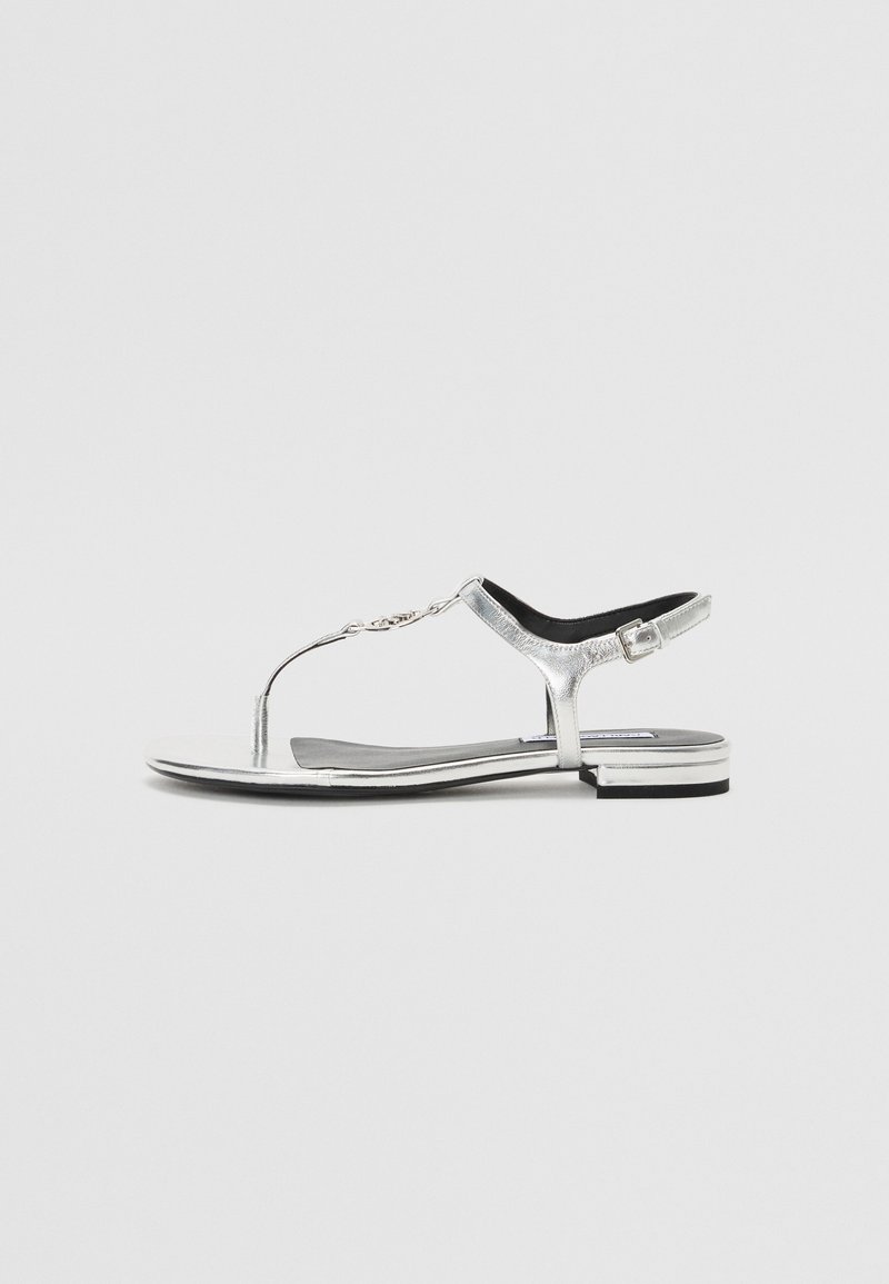 Sølv flate thong-sandaler med T-stropp-design, med kjedeakcent, glatt metallisk overflate, svart polstret såle og lav hæl.