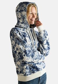 Młoda osoba uśmiechająca się, ubrana w białą bluzę z kapturem z niebieskim motywem przyrody i zwierząt, z rękami schowanymi w rękawach oraz w niebieskie dżinsy.