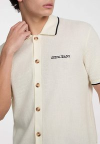 Cremfarbener Poloshirt mit strukturiertem Gewebe, ausgestattet mit einem kontrastierenden schwarzen Kragen, goldenen Knöpfen und gesticktem "GUESS JEANS"-Logo.