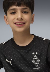 Ragazzo sorridente che indossa una maglia da calcio nera del Borussia Mönchengladbach con il logo Puma e il testo "125 Jahre" su uno sfondo grigio.