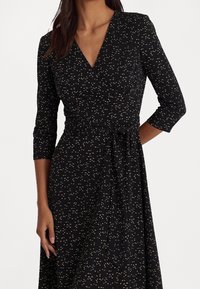 Svart omlottklänning med trekvartslånga ärmar, en knytning i midjan och ett spritt mönster av guld och krämfärgad i hela plagget. Slät tygtextur.