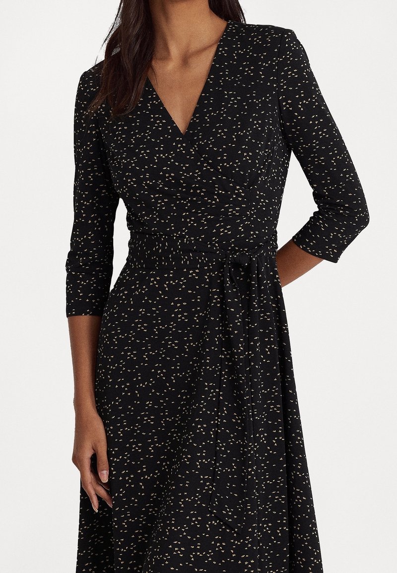 Svart omlottklänning med trekvartslånga ärmar, en knytning i midjan och ett spritt mönster av guld och krämfärgad i hela plagget. Slät tygtextur.