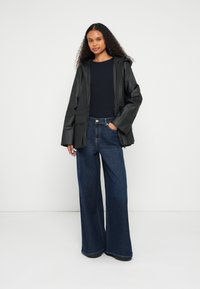 Μαύρο αδιάβροχο μπουφάν με κουκούλα, φορεμένο πάνω από μπλε σκούρο τοπ και άνετα φαρδιά μπλε τζιν παντελόνια, με κουμπί και τσέπες.