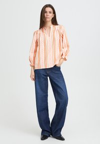 Femme portant un chemisier rayé orange et blanc à manches longues, avec un jean large bleu foncé, se tenant avec une main dans la poche.