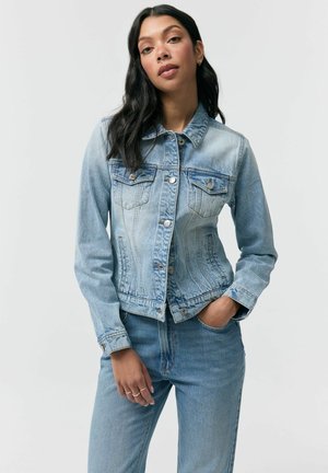 Młoda kobieta w jasnoniebieskiej dżinsowej kurtce i dżinsach, stojąca z jedną ręką w kieszeni na jednolitym tle.