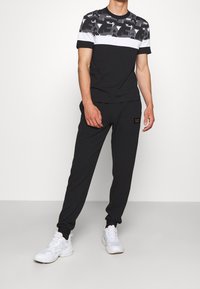 Completo sportivo bianco e nero composto da una maglietta a maniche corte con motivo e una striscia bianca, abbinata a pantaloni jogger neri con una patch del logo.