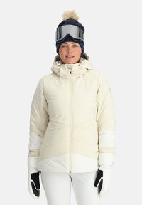 Weiße isolierte Skijacke mit Zickzackmuster, verstellbarer Kapuze und Reißverschlusstaschen. Modell trägt eine marineblaue Strickmütze und schwarze Handschuhe.