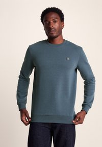 Dark teal sweatshirt made of soft fabric, featuring a crew neck, ribbed cuffs, and a small logo on the left chest. Worn with dark jeans.