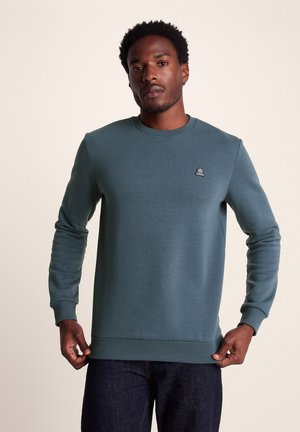 Dark teal sweatshirt made of soft fabric, featuring a crew neck, ribbed cuffs, and a small logo on the left chest. Worn with dark jeans.