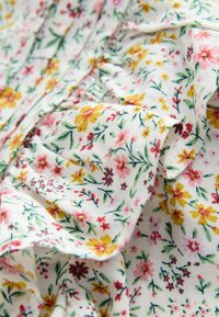 Blumenstoff mit kleinen, bunt gemischten Blumen in Pink, Gelb und Lila auf einem weißen Hintergrund, mit sanften Falten und Textur.