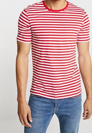 Uomo che indossa una maglietta a righe orizzontali rosse e bianche a maniche corte e jeans blu, in piedi davanti a uno sfondo chiaro e uniforme.