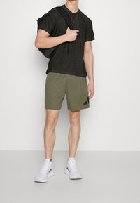 Černá krátká halenka s texturovaným vzorem, olivově zelené šortky s černým akcentem a bílé sportovní boty. Batoh přes jedno rameno.